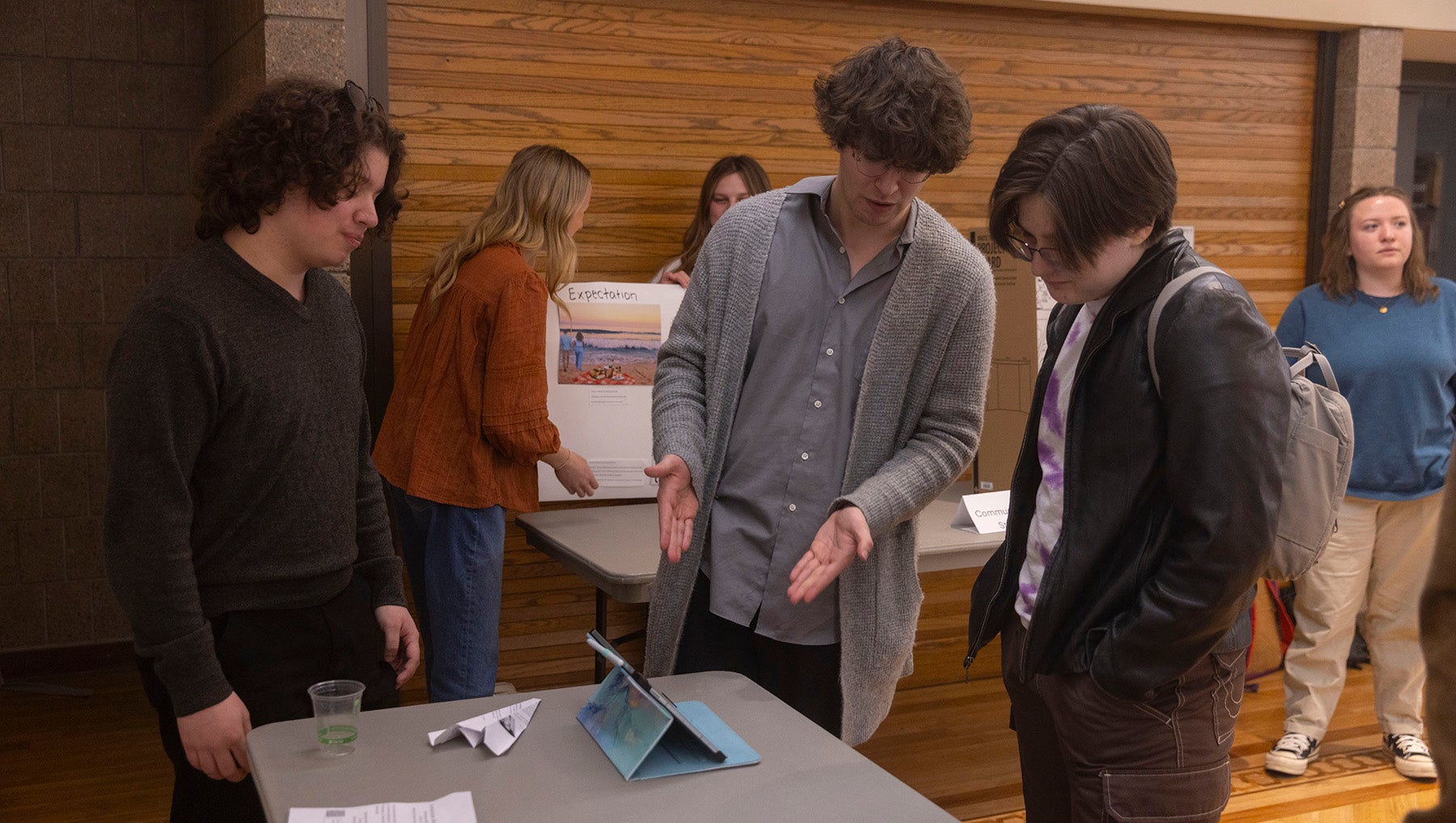 Students congregate around a table to review a communications presentation on a digital tablet.