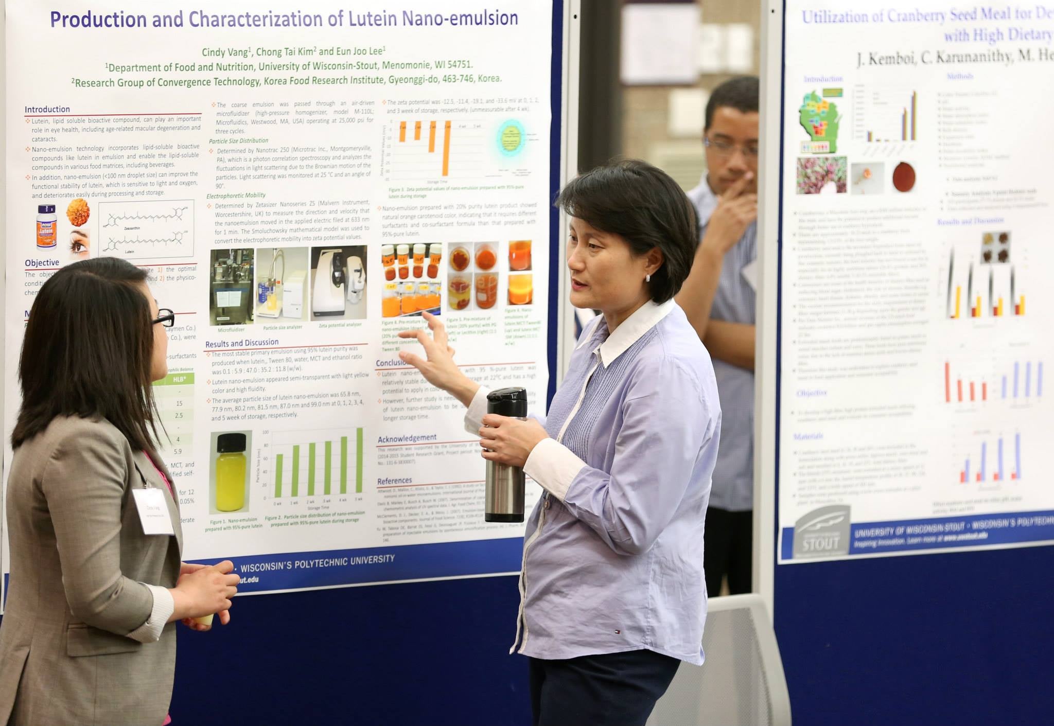 Two people stand in front of a research poster titled “Production and Characterization of Lutein Nano-emulsion.” One person gestures toward the poster while holding a tumbler, and the other listens. Additional research posters are displayed in the background.