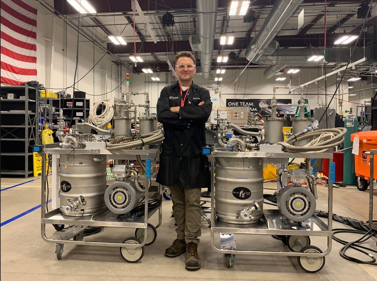 A person stands with arms crossed between two stainless steel vacuum cart ground support equipment units on wheeled frames inside an industrial aerospace facility. Each cart has hoses, valves and gauges, with high ceilings, ductwork and an American flag visible in the background.