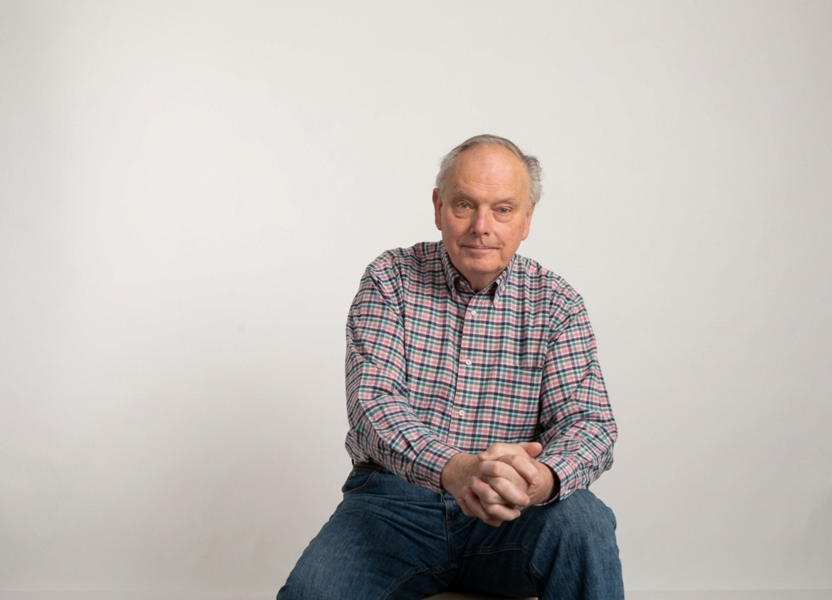 A person sits facing the camera against a light background, wearing a plaid button-down shirt with hands clasped.