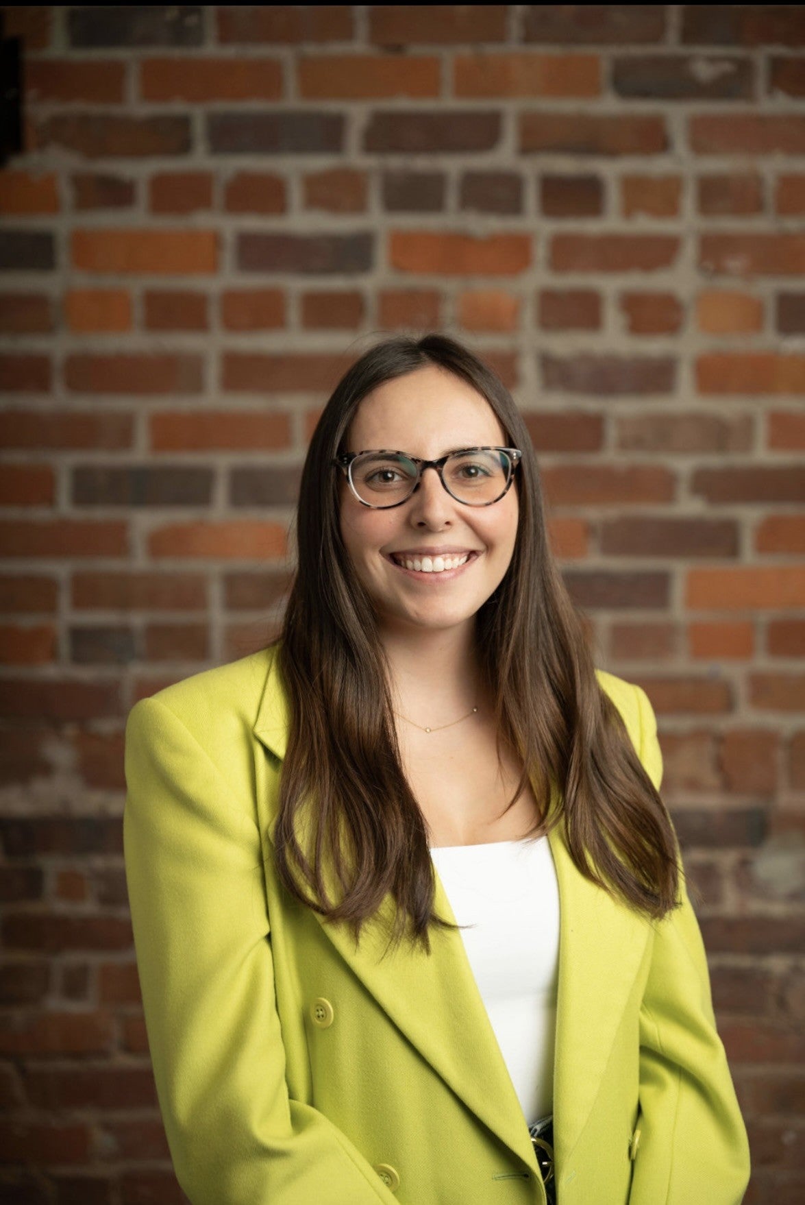 Professional headshot of a person with long dark hair wearing glasses, a bright green blazer, and a white top, standing in front of a brick wall and facing the camera.