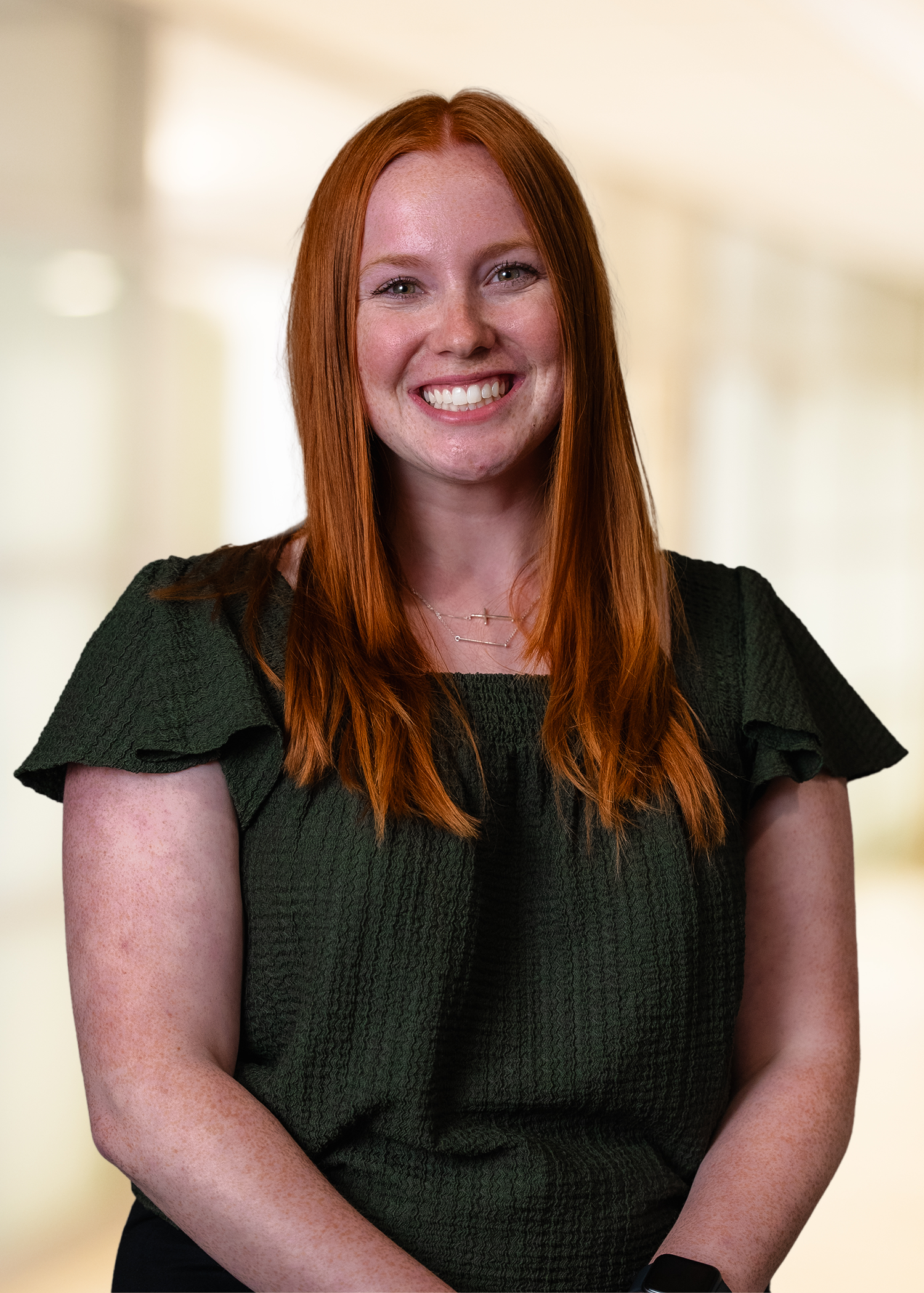 Person with long red hair smiling at the camera, seated against a softly blurred indoor background.
