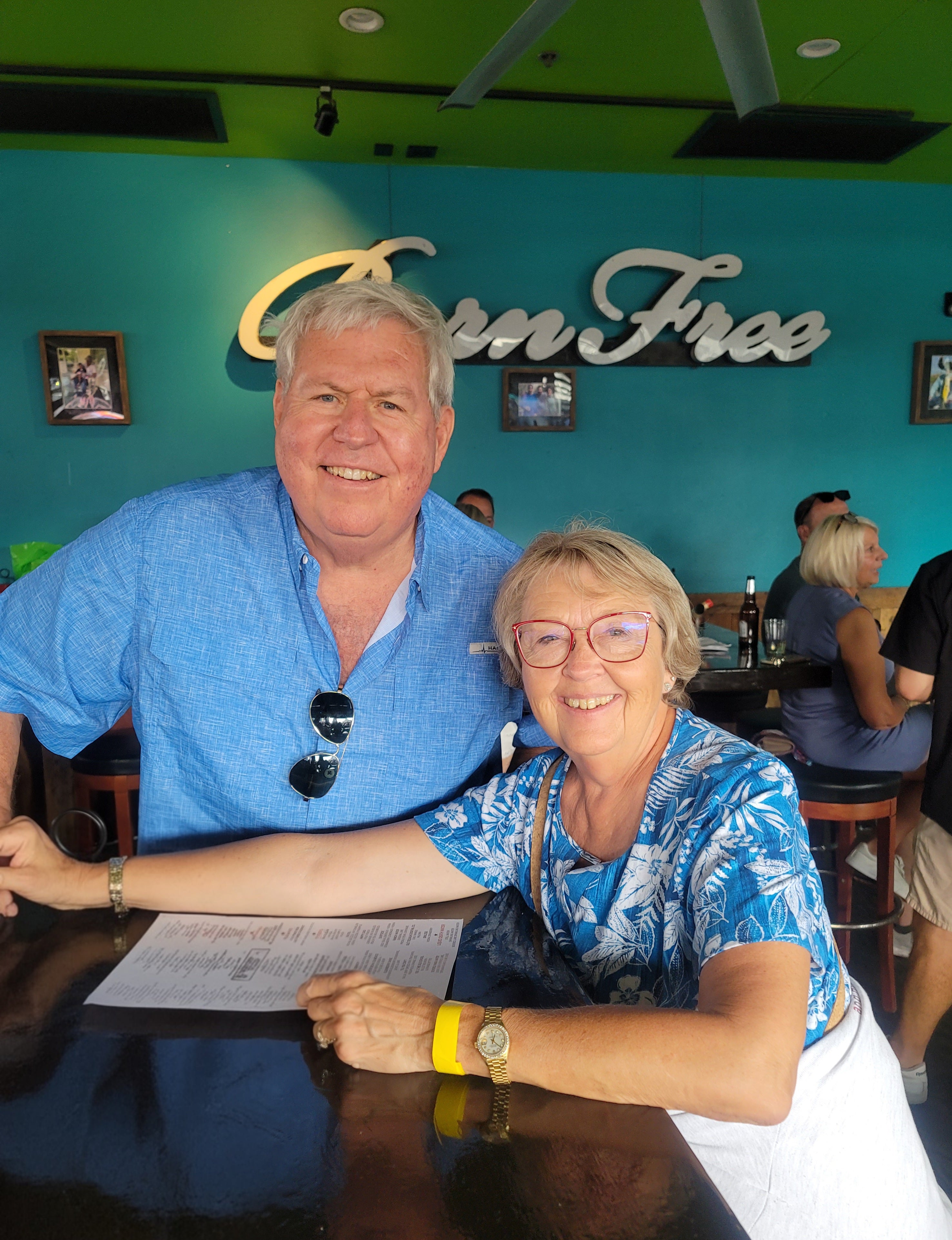 Two older adults seated at a restaurant table, smiling at the camera, with a turquoise wall and “Green Tree” sign behind them.