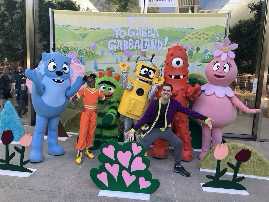 Michael Hilliger poses with costumed characters from Yo Gabba GabbaLand! in front of a promotional display featuring flowers and a colorful backdrop.