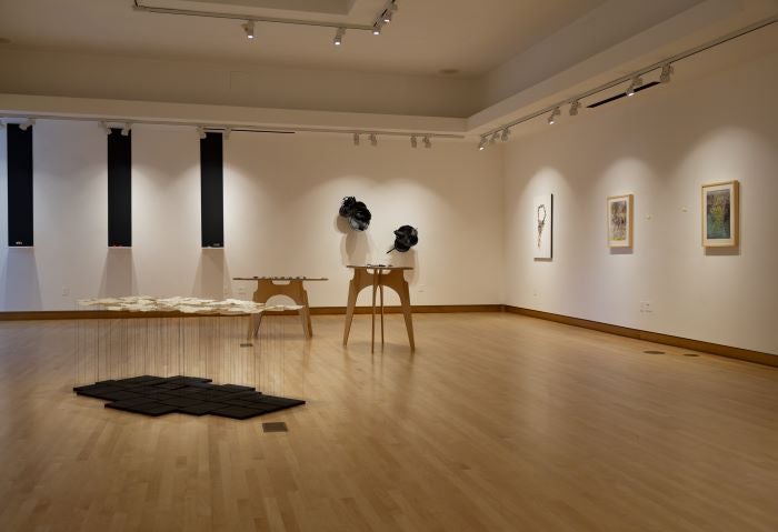 Two walls of various art pieces, with two tables in the center of a space displaying smaller pieces.