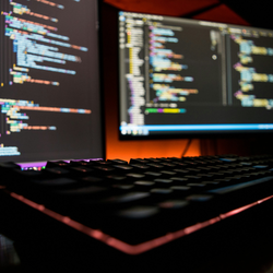 Close up of a black computer keyboard with a red glow underneath. In the blurry background, two computer monitors are showing lines of code on dark screens.