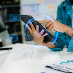 A close-up of a person's hands interacting with a black tablet at a white desk. Glowing blue, futuristic digital icons and a central circular graphic containing the letters "AI" are superimposed over the device and surrounding space.