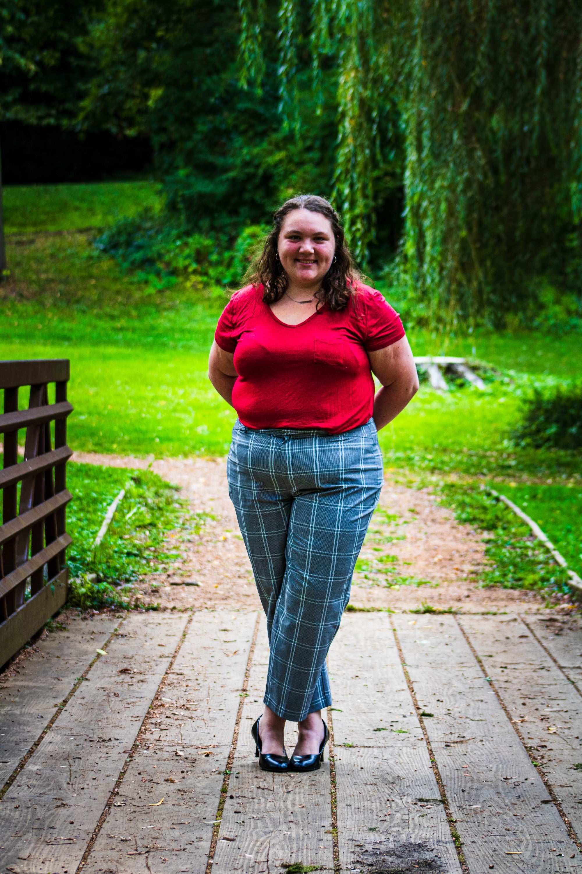 Alisha Marquardt is a white woman with shoulder length wavy brown hair. She is standing on a bridge outside wearing a red shirt and gray pants.