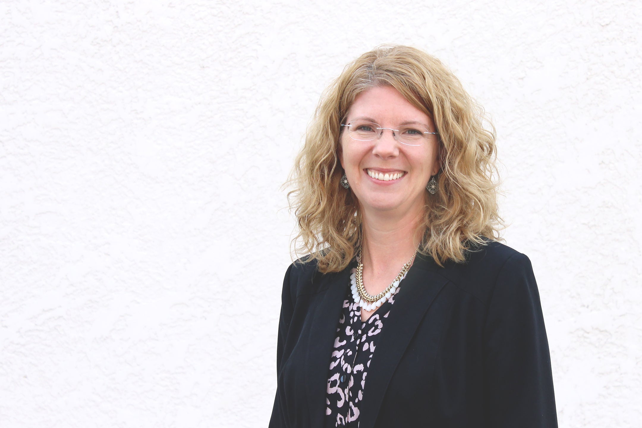 Brenda Scheurer is a white woman with curly shoulder length blonde hair and glasses. She is wearing a black blazer and printed black shirt and is standing in front of a white backgorund. 