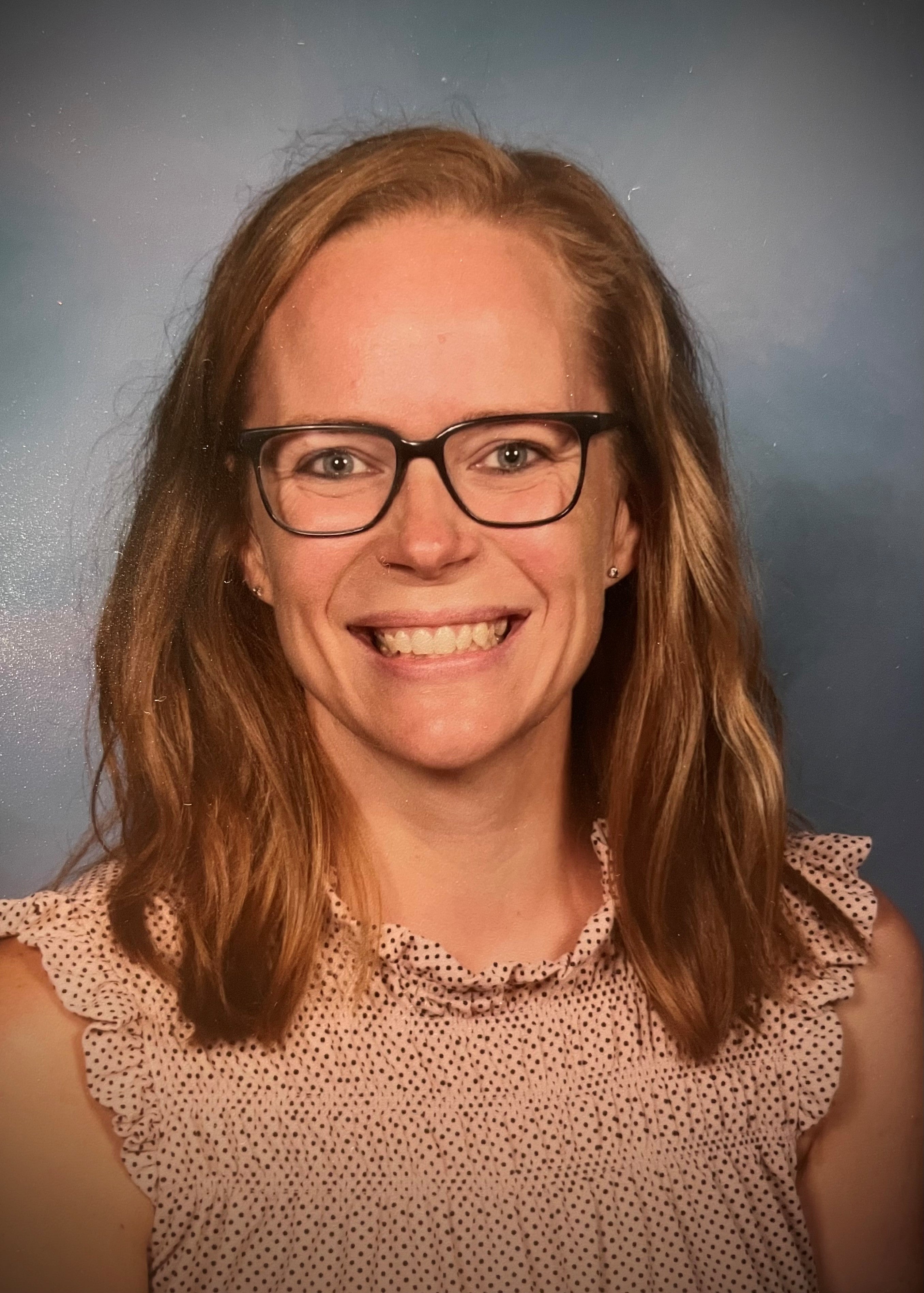 Carly Quick is a white woman with dirty blonde shoulder length hair. She is wearing glasses and a polka dotted shirt.