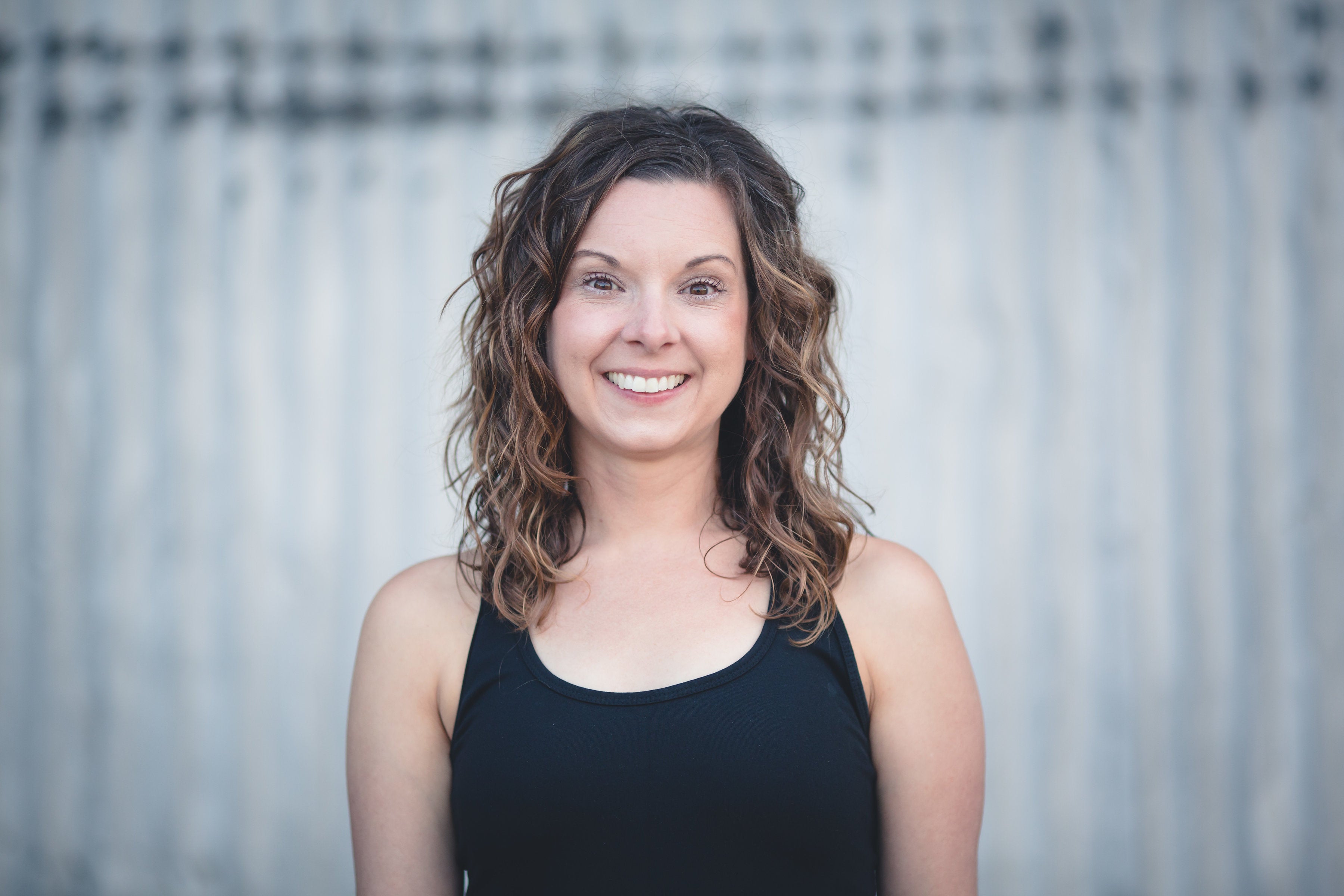 Christy Langman is a white woman with shoulder length, curly hair. She is wearing a black tank top and standing in front of a gray background.