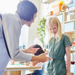 An instructor kneels to engage with a young student in a bright, classroom-like setting with art supplies and shelving visible in the background.