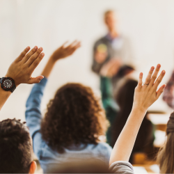 Students raising their hands in a classroom, with an instructor visible in the background.