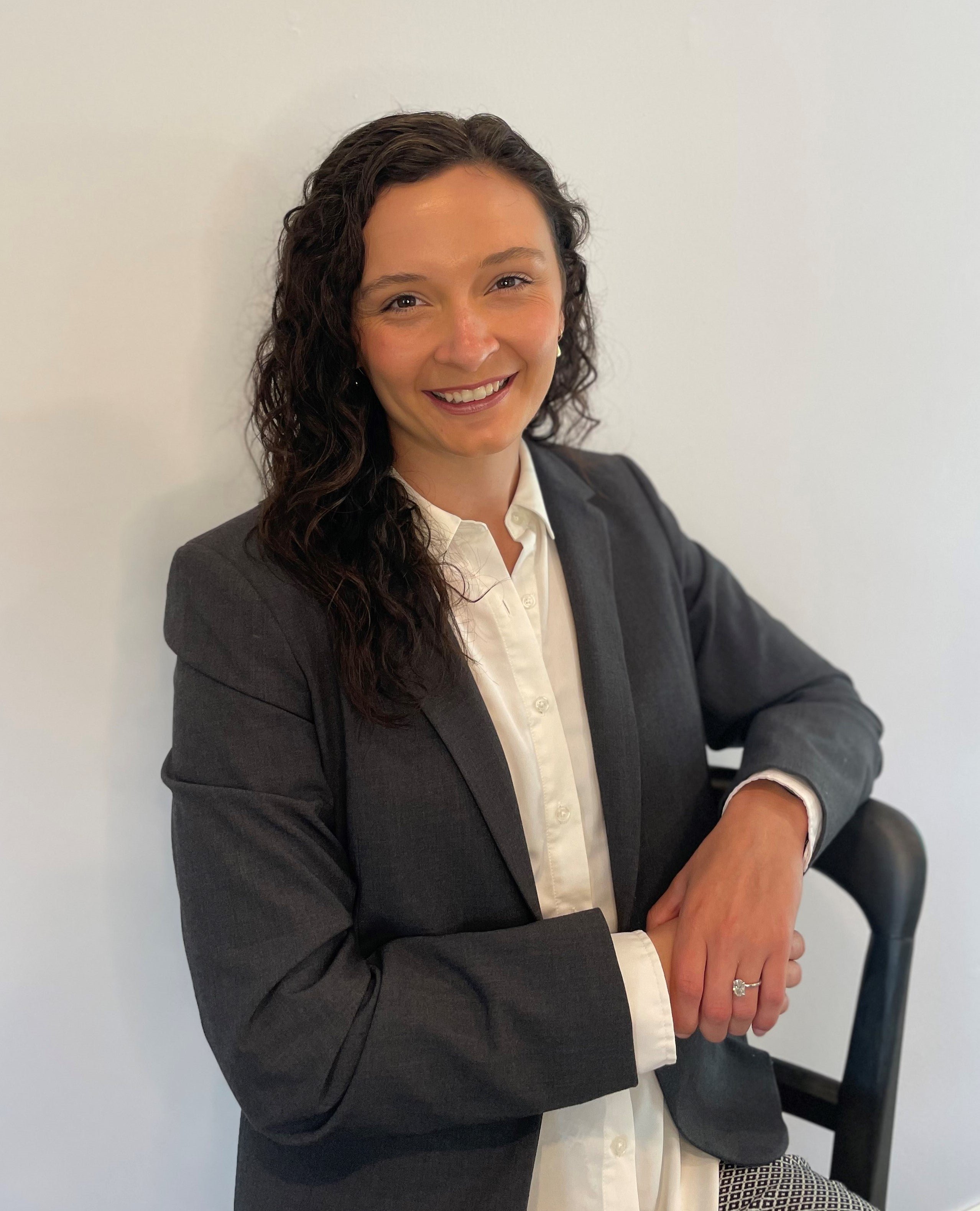 Erica Premeu is a tan woman with long, curly black hair. She is wearing a white shirt and gray blazer in front of a white background.