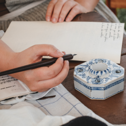 A person handwriting in a notebook beside a delicate blue and white ceramic ink holder, evoking the personal and reflective nature of memoir writing