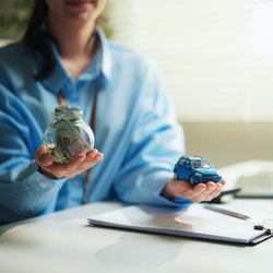 A person wearing a light blue button-down shirt sits at a white desk. They are holding a small glass jar filled with folded money in their right hand and a small blue toy car in their left hand. A clipboard with paper and a pen are resting on the desk in front of them.