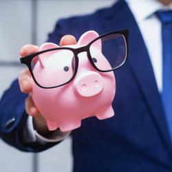 A person wearing a dark blue suit and a blue tie is holding up a pink piggy bank. The piggy bank is wearing black-rimmed eyeglasses.