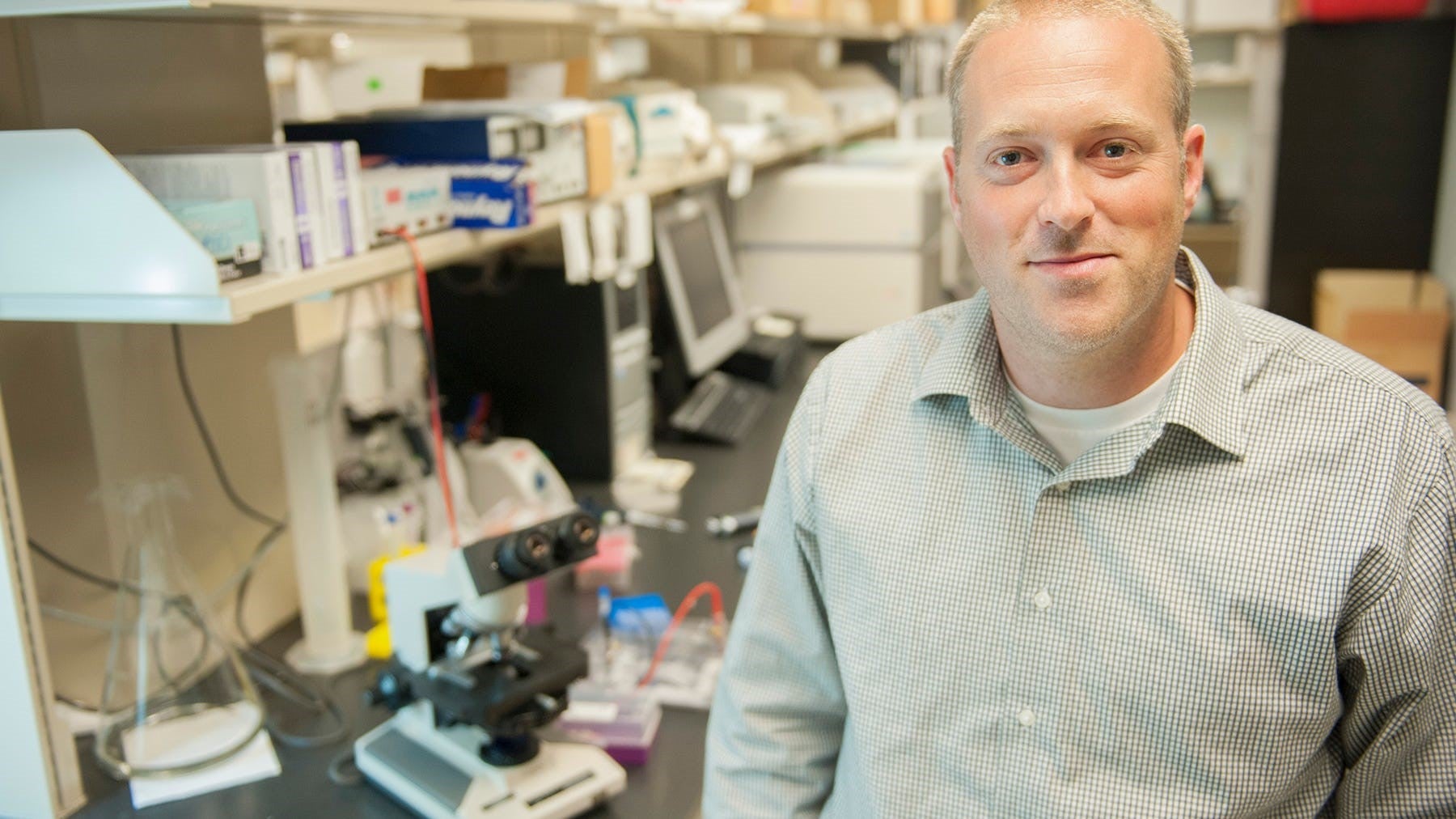 Dr. Scott Hebbering is a white man with short blond hair and a light gray polo shirt. He is sitting in a laboratory desk.