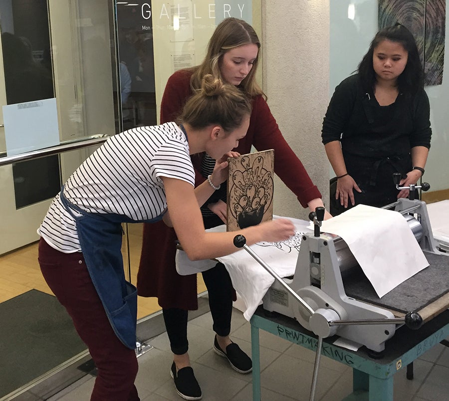 UW-Stout students Kendra Lundstrom, Victoria Pesch and Tracie Newman remove a wooden relief plate after printing a t-shirt