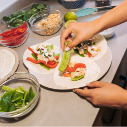 Alt text:  "Hands assembling tacos on white tortillas, topped with shrimp, sliced red peppers, avocado, and fresh herbs, surrounded by bowls of colorful ingredients on a counter.