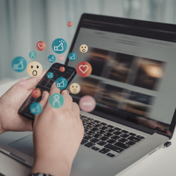 A person holds a smartphone with colorful social media icons floating above it, including emoji reactions, a heart, and engagement symbols, with a laptop open in the background.