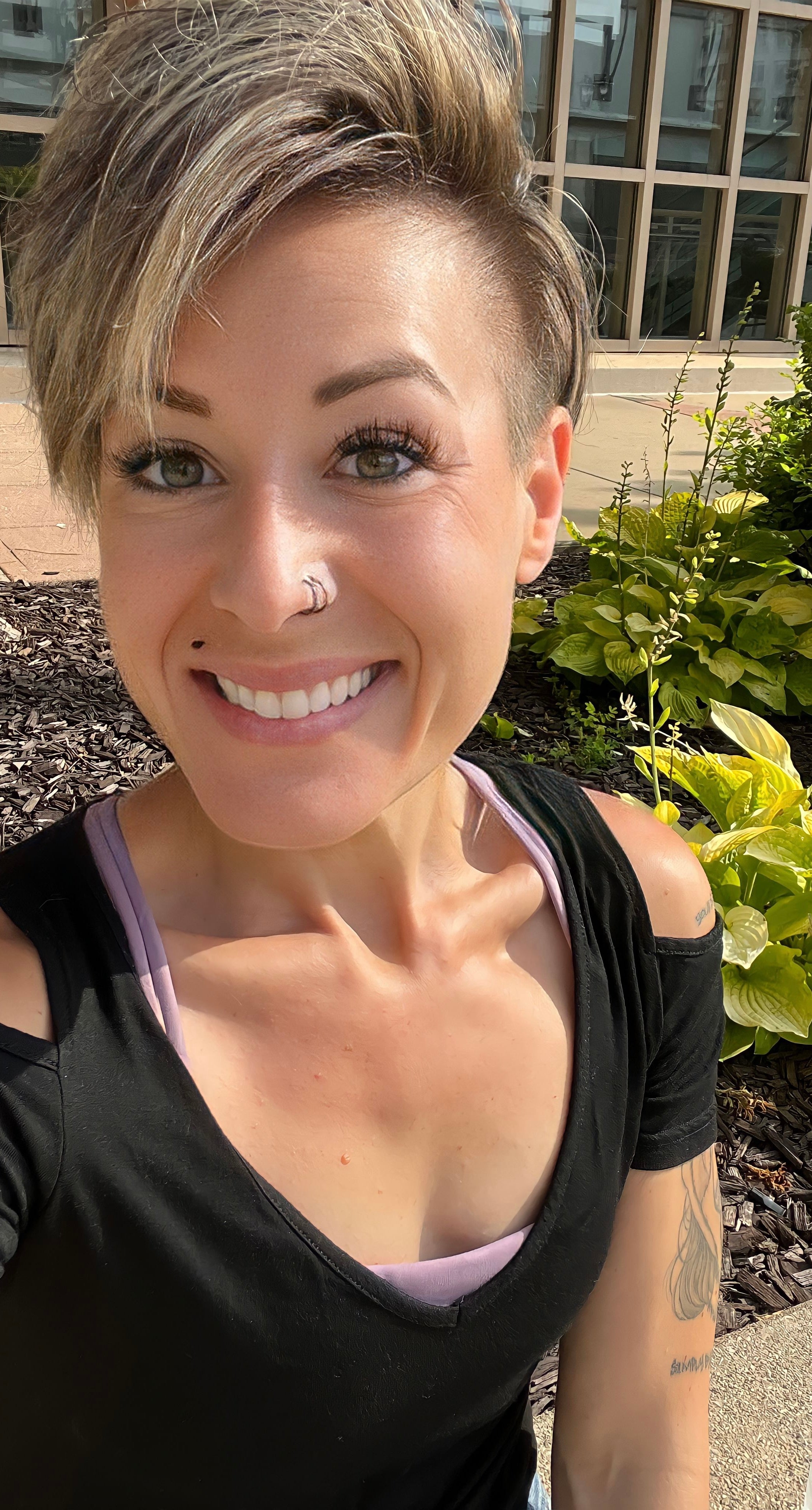 Teresa Hand is a white woman with short dirty blonde hair and a nose ring. She is wearing a black cold shoulder top and standing near a garden.