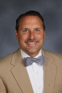 Tom Cardamone is a white man with short hair and a short beard and mustache. He is wearing a brown suit jacket and gray bow tie.