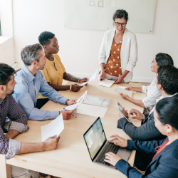 A team of 7 people sitting around, with a woman standing up