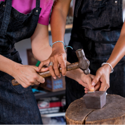 Two people hammering metal on an anvil during a metalworking or jewelry-making workshop.