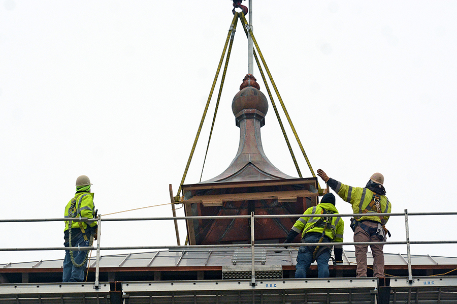 The cupula is maneuvered into place by a crane operator and workers on top of Bowman Hall. The cupula had to fit over a metal pole and then be fastened to the roof.