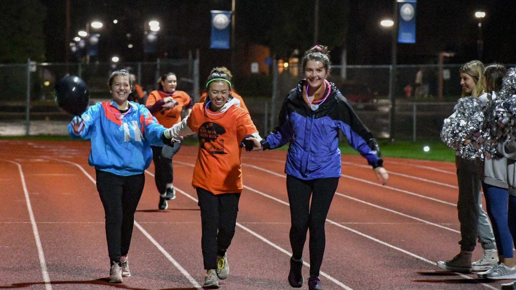 Three girls running on a track, hands linked, and smiling.