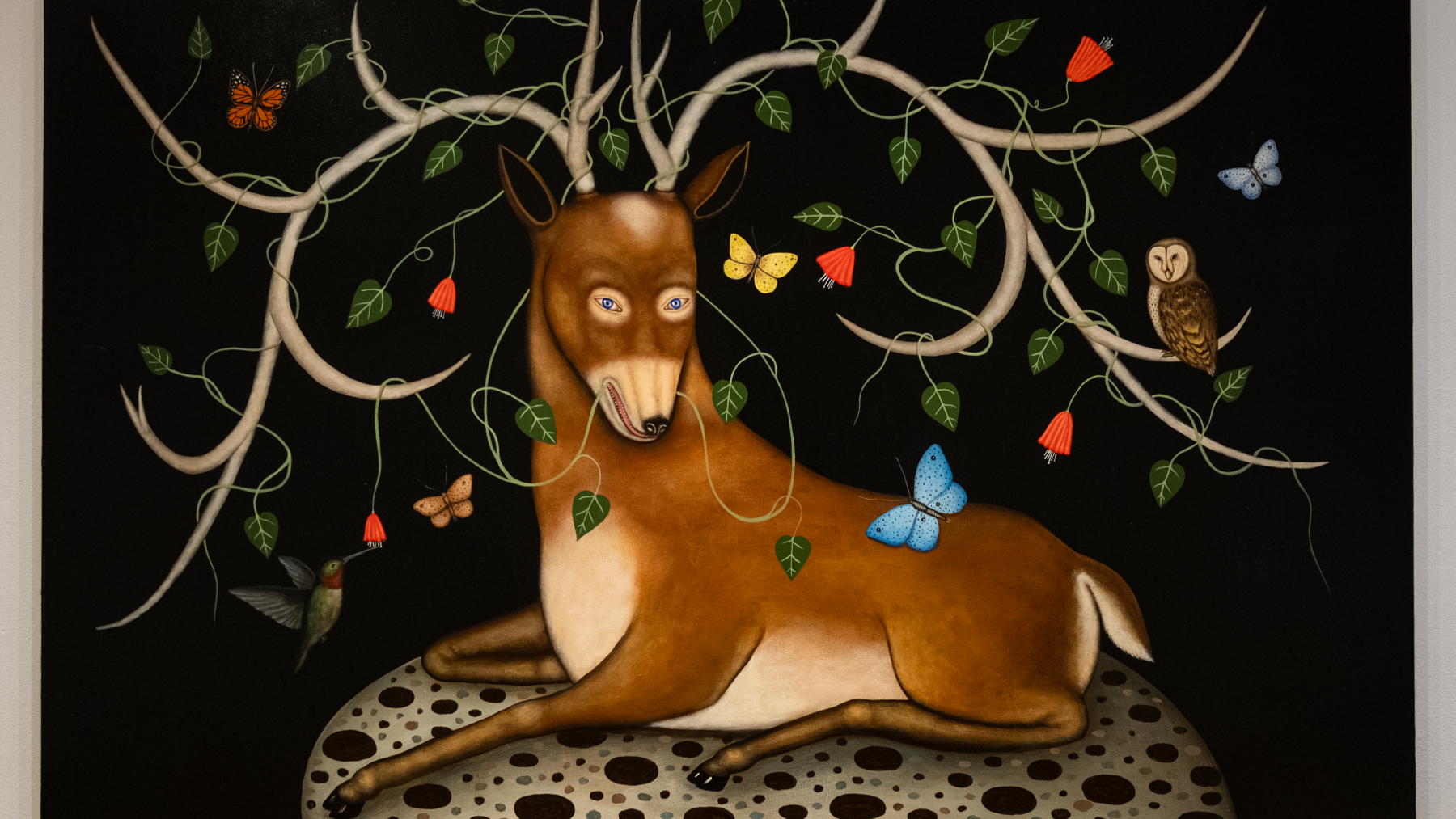 Acrylic painting captures a Stag centered siting on rock bed with large antlers. A vine coming out of the deer's mouth circles the antlers, and butterflies, an owl and a humming bird interact with the composition. Flying around the deer or sitting on an antler.  