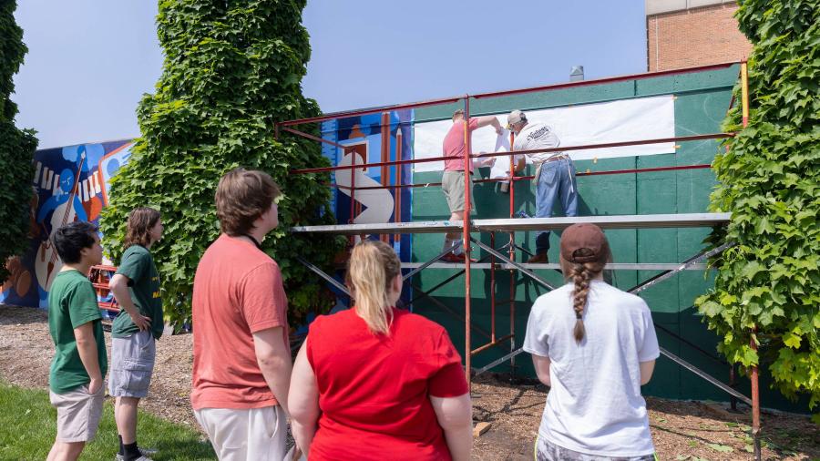 Students watch as other students stand on scaffolding to trace the top half of a tall mural.