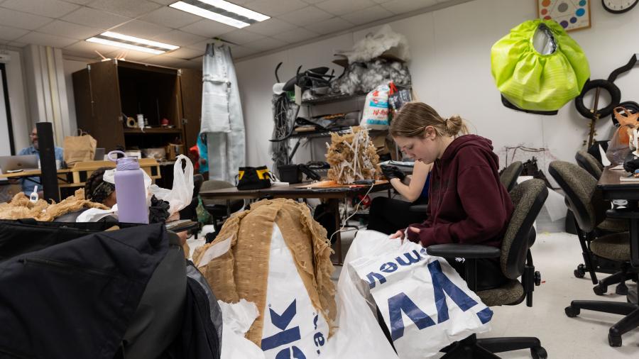 A student sits at a table amongst many materials all around them, on the floor and on the table.