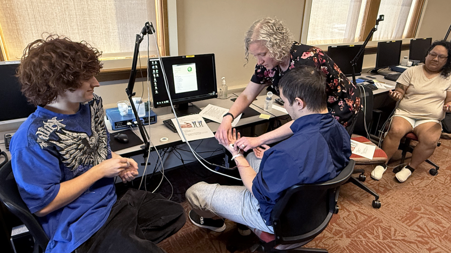 Two students sit at a desk outfitted with sensory testing equipment while a professor explains a psychology experiment.