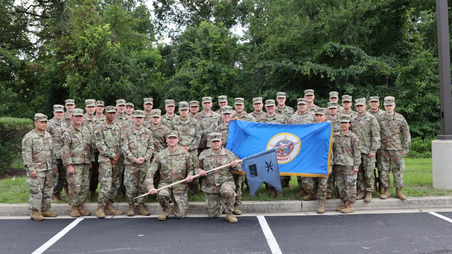 Lieutenant Colonel Brian Morgan stands with a large group of Minnesota National Guard soldiers in uniform outdoors, with the Minnesota state flag displayed at the center of the group.
