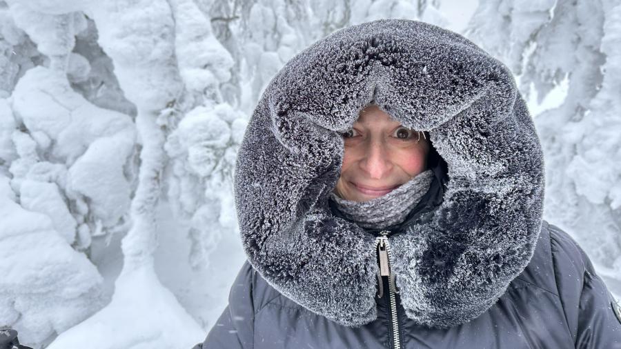 A person bundled in heavy winter clothing stands in a snowy forest, surrounded by trees completely covered in thick frost and snow. Their large fur-lined hood is coated in ice crystals, indicating extremely cold weather conditions.