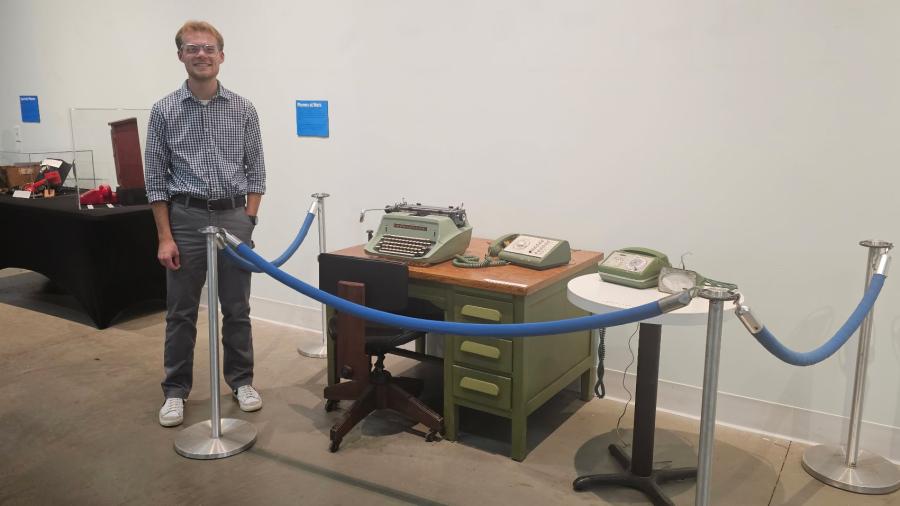 A person stands behind a blue rope barrier in front of a historical office setup that includes a green metal desk, a vintage typewriter, and several rotary telephones. Additional display items sit on a nearby black table.