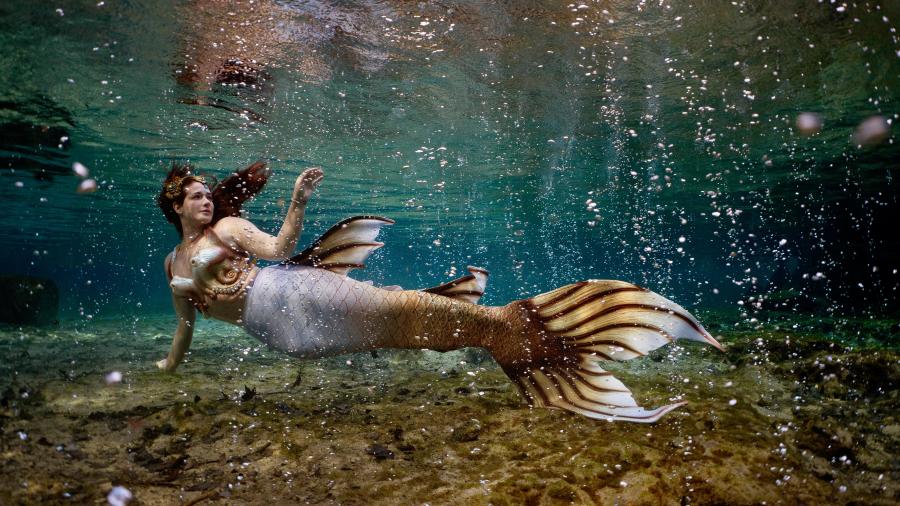A woman dressed as a mermaid swims underwater in clear green-blue water, her long hair floating around her as bubbles rise to the surface above a rocky streambed.