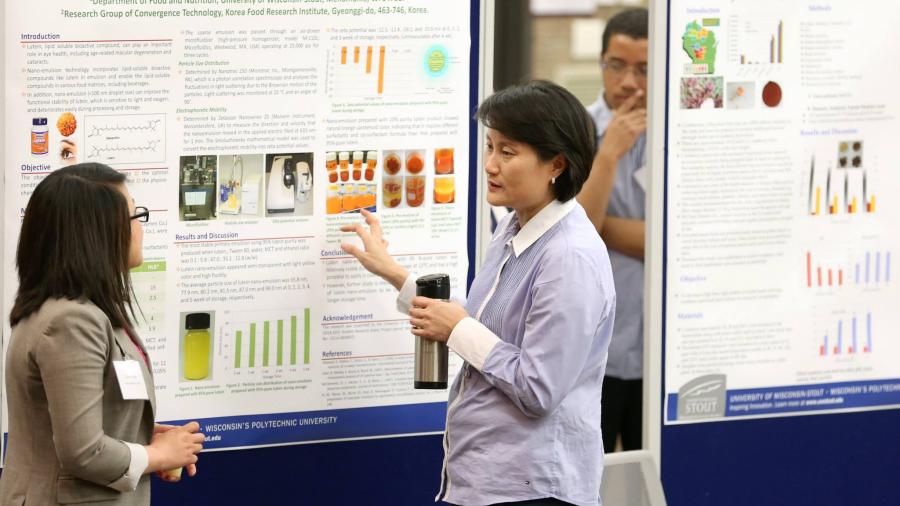 Two people stand in front of a research poster titled “Production and Characterization of Lutein Nano-emulsion.” One person gestures toward the poster while holding a tumbler, and the other listens. Additional research posters are displayed in the background.