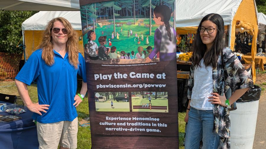 Two people stand on either side of a tall “Powwow Bound” promotional banner at an outdoor event with vendor tents and trees in the background.