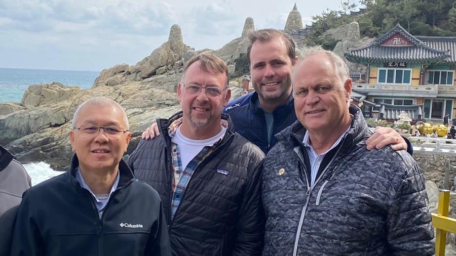 Four people stand together on a coastal walkway with rocky cliffs and a traditional-style building in the background.