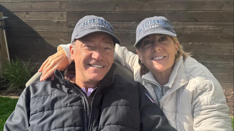 Two adults wearing “Blue Devils Alumni” hats sit outdoors on a bench, leaning together for a close-up photo with a wooden fence and greenery behind them.
