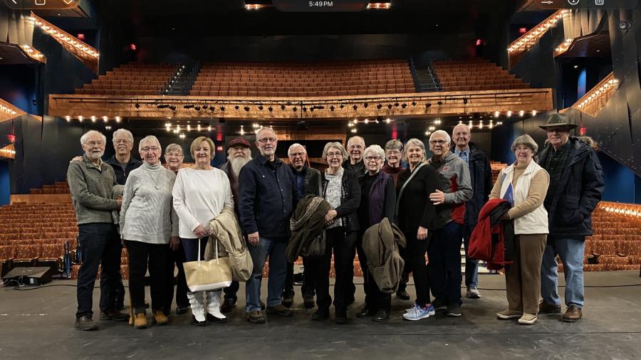A group of visitors stands on a theater stage in front of rows of auditorium seating and a balcony, posing for a photo during a venue tour. Warm stage and balcony lights illuminate the theater interior.  Provide your feedback on BizChat