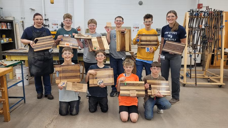 Students and instructors stand in a woodworking shop displaying completed cutting boards, trays and other wood projects made during a STEAM construction camp.