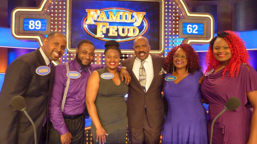 Group of six people stand together on the Family Feud game show set with host Steve Harvey. The illuminated Family Feud scoreboard is visible behind them.