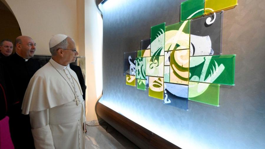 A religious leader in white robes views a large illuminated artwork composed of green, yellow and blue translucent panels.