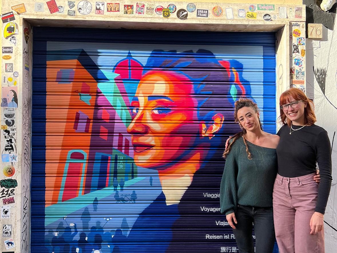 Two people stand in front of a mural of the portrait of a women