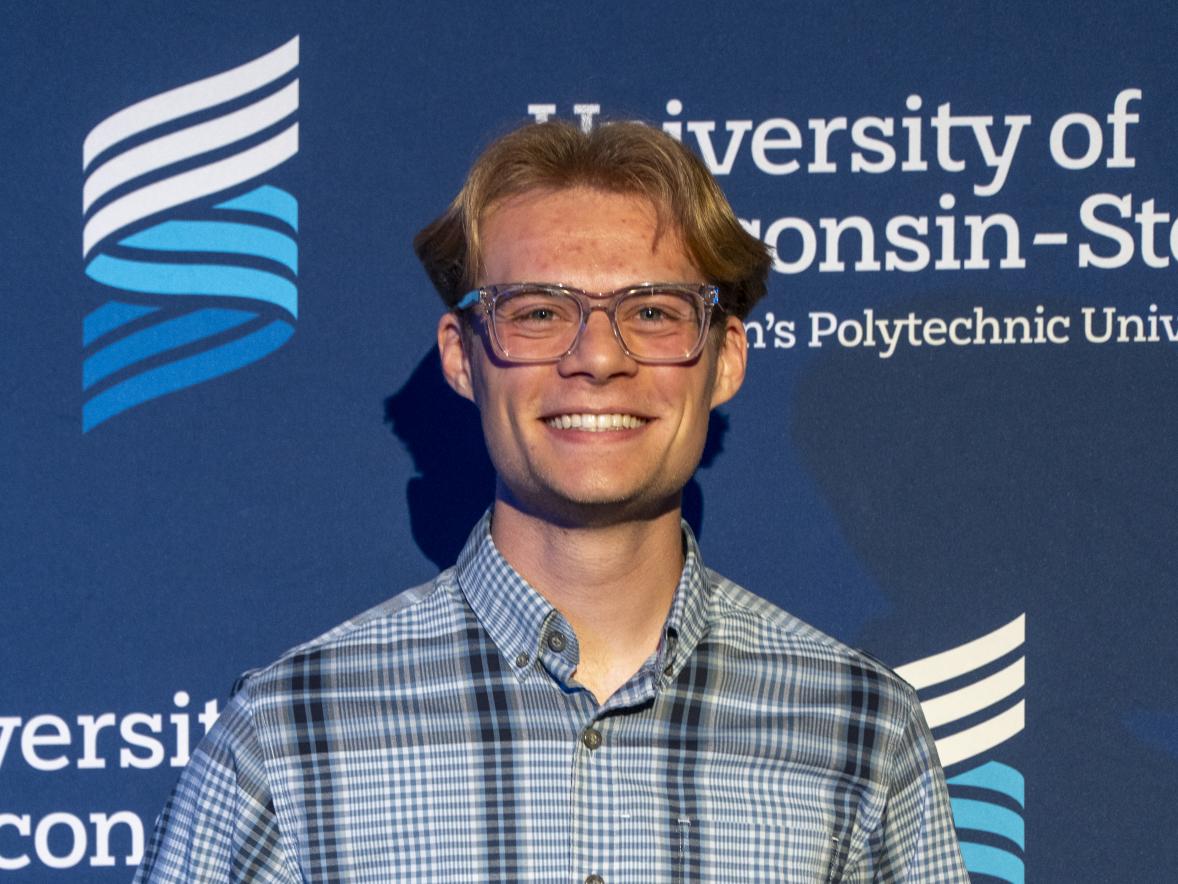 A person stands in front of an official University of Wisconsin–Stout backdrop while holding a clear award plaque. They are wearing a plaid shirt and slacks.