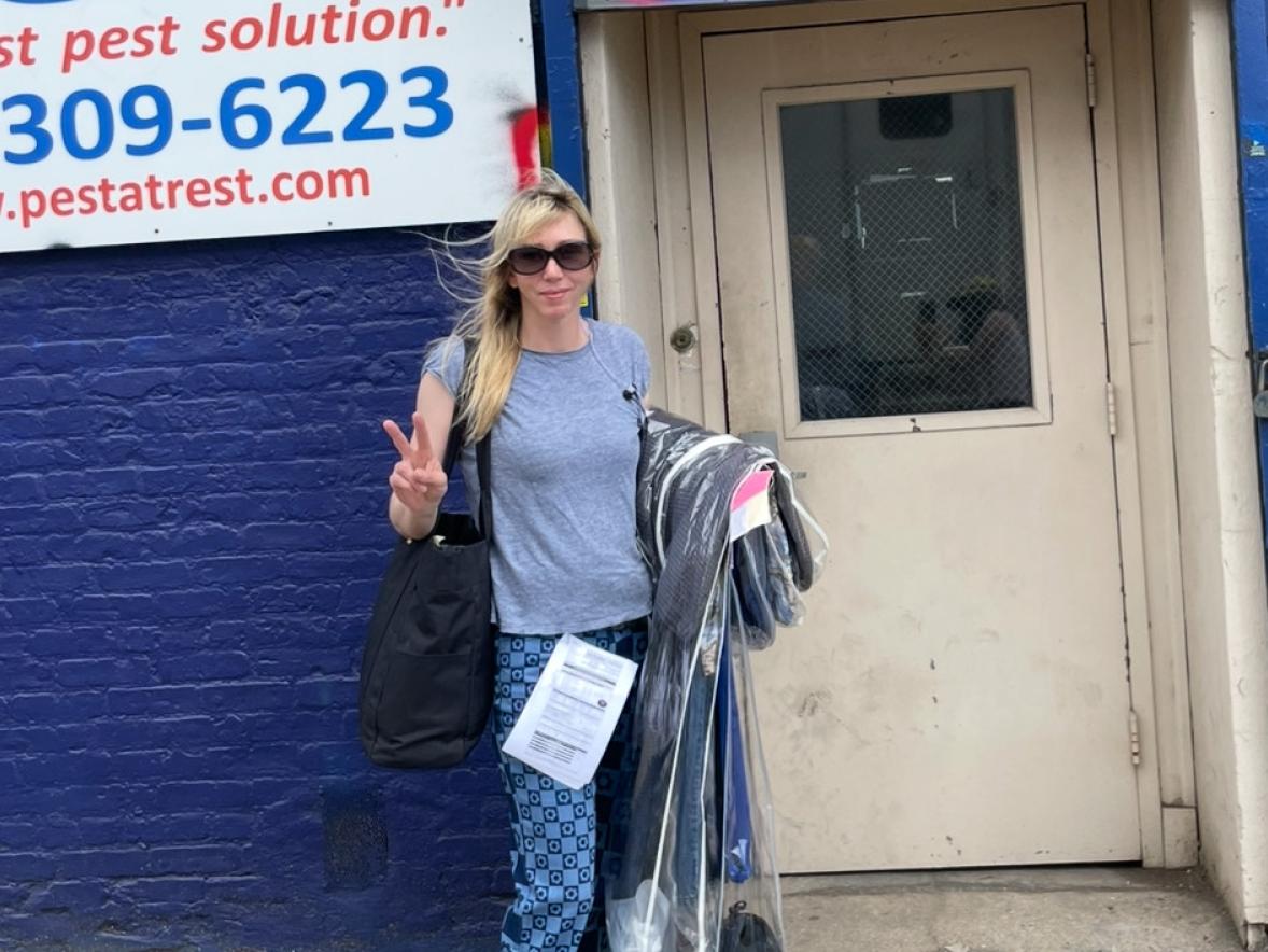 A person is standing on a sidewalk in front of a blue brick wall and a beige door.  The person is wearing a gray T-shirt, patterned blue pants, and black-and-white sneakers. They are holding a black shoulder bag and carrying several clothing items on hangers covered in clear plastic.  piece of paper is tucked into the waistband of the pants, and the person is making a peace sign with one hand.